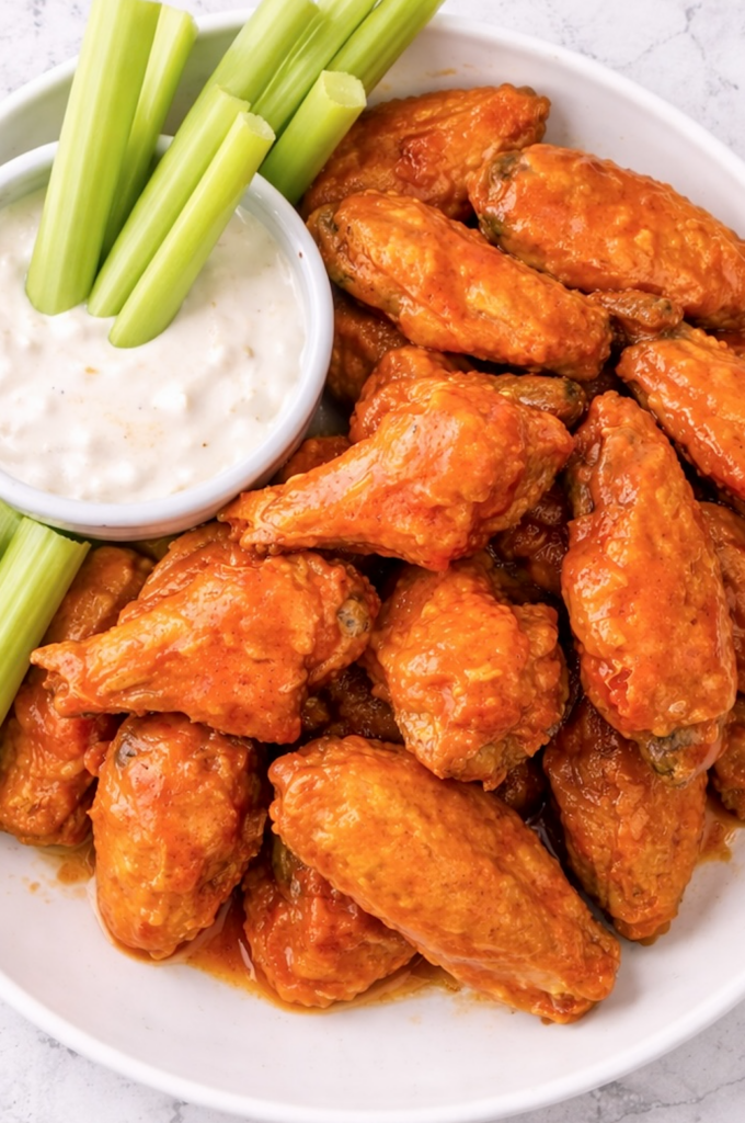 Crispy buffalo chicken wings coated in tangy sauce with celery sticks and blue cheese dressing on a white plate.