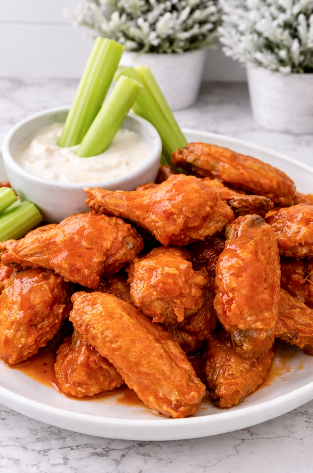 Alt text: Oven-baked buffalo chicken wings piled on a plate with celery sticks and blue cheese dip in the background.