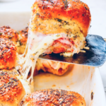 Ham and cheese Hawaiian roll sliders being lifted from a baking dish, showing melted Swiss cheese stretching between layers of ham and soft rolls.