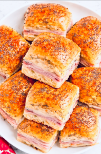 Stack of baked ham and cheese Hawaiian roll sliders on a white plate, brushed with butter and topped with poppy seeds.