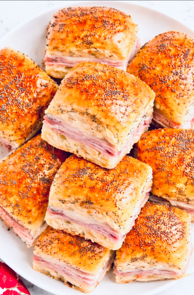 Stack of baked ham and cheese Hawaiian roll sliders on a white plate, brushed with butter and topped with poppy seeds.