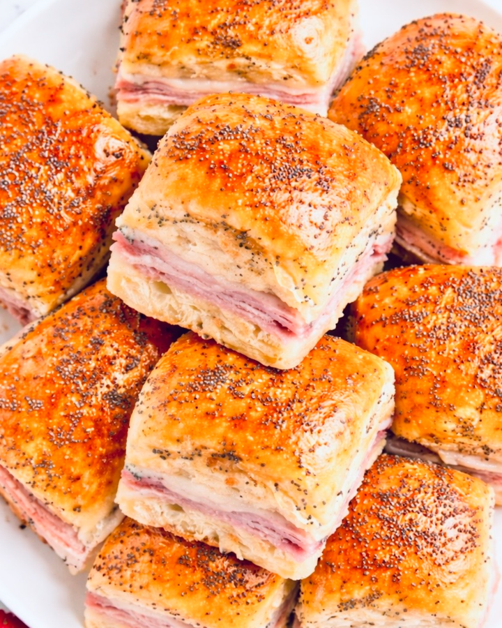 Stack of baked ham and cheese Hawaiian roll sliders on a white plate, brushed with butter and topped with poppy seeds.