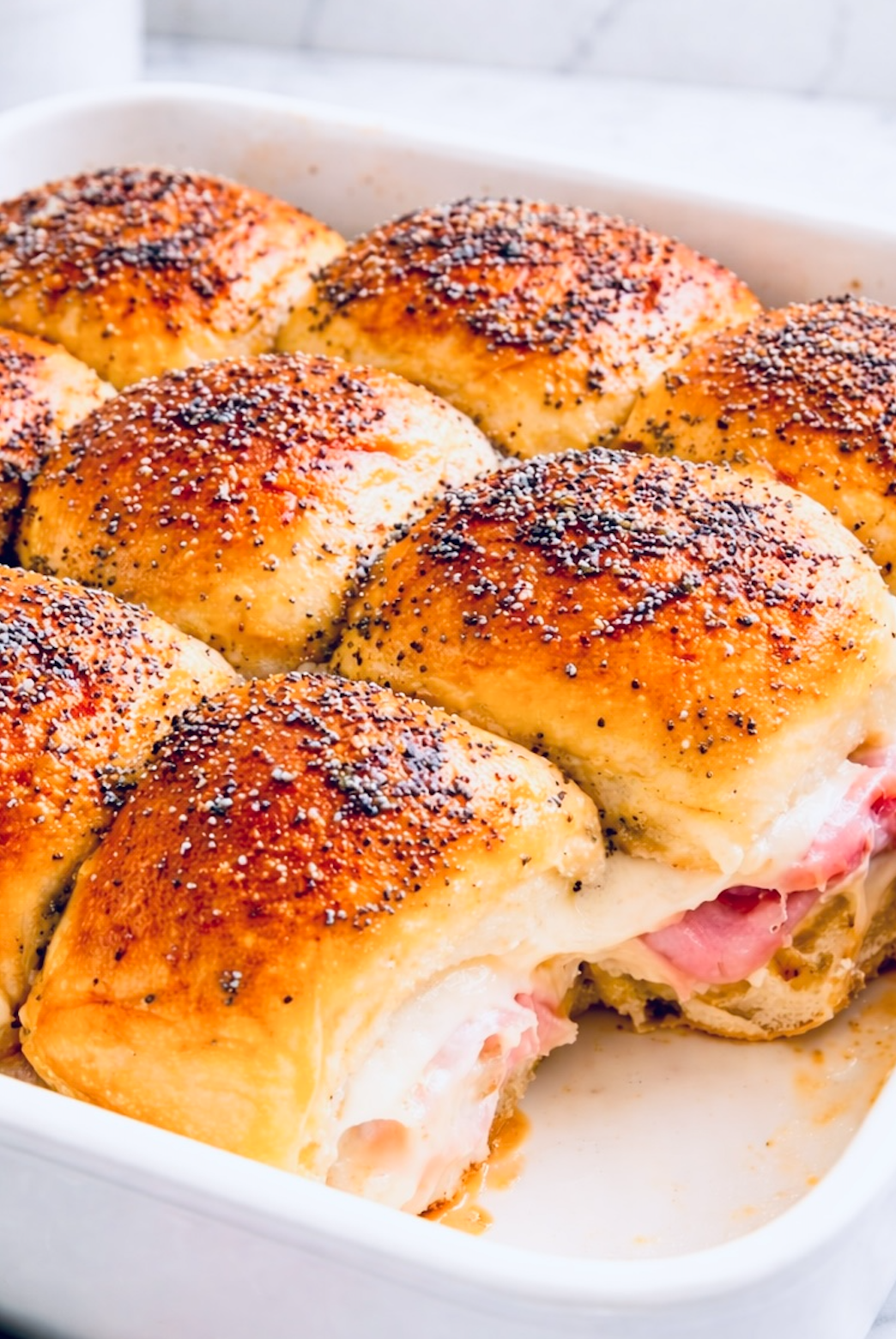 Ham and cheese Hawaiian roll sliders baked in a white casserole dish, topped with butter and poppy seeds.