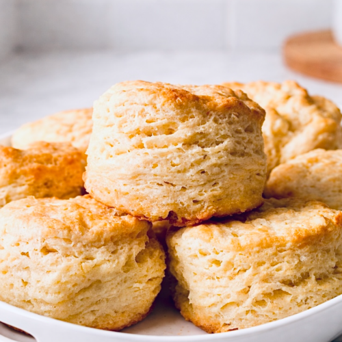 Close-up of tall whipping cream biscuits with flaky layers and golden brown tops stacked in a white baking dish.
