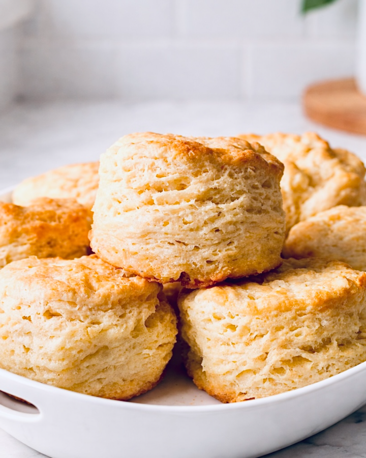 Close-up of tall whipping cream biscuits with flaky layers and golden brown tops stacked in a white baking dish.