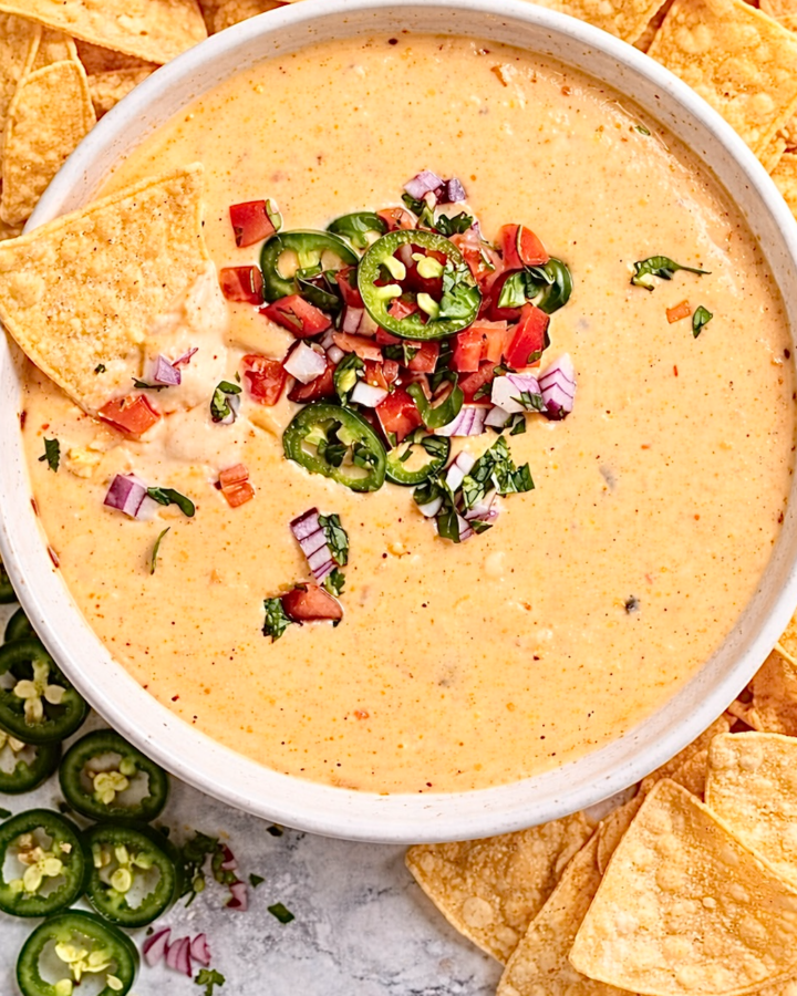 Bowl of creamy Rotel cheese dip topped with chopped tomatoes, jalapeño slices, red onion, and cilantro, surrounded by tortilla chips.