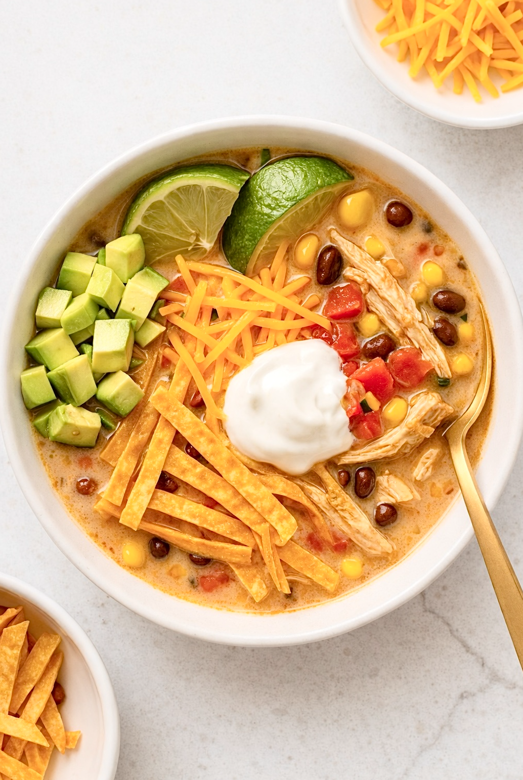 Bowl of creamy chicken tortilla soup topped with avocado, shredded cheese, tortilla strips, sour cream, and lime wedges on a light countertop