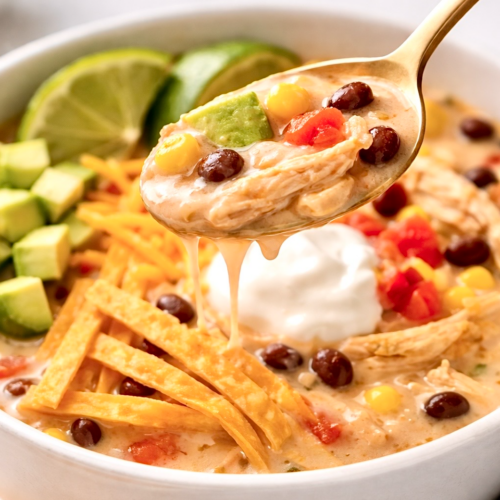 Spoon lifting creamy chicken tortilla soup showing shredded chicken, black beans, corn, avocado, and melted cheese in a rich broth