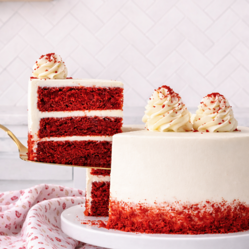 Three-layer red velvet cake with cream cheese frosting, shown with one slice lifted to reveal bright red cake layers and white frosting.