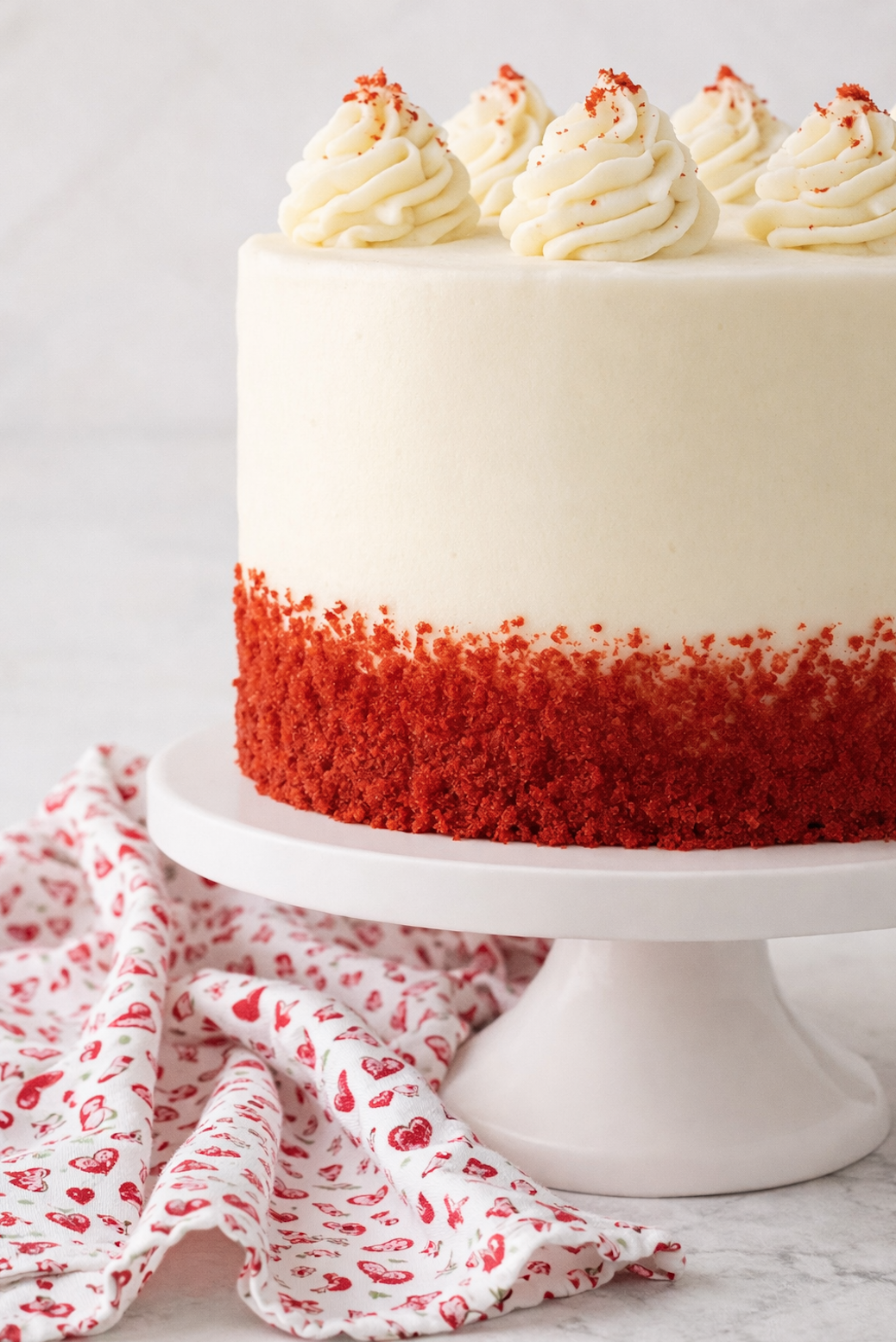 Frosted red velvet cake on a white cake stand, finished with cream cheese frosting and red cake crumbs around the base.