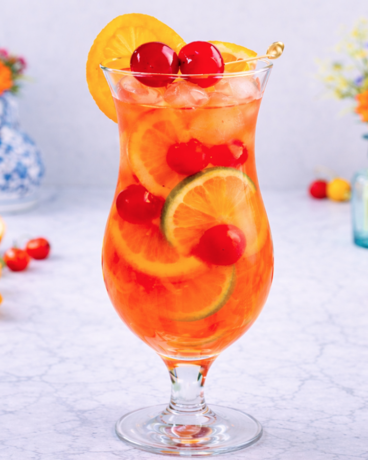 Single hurricane cocktail in a clear hurricane glass with ice, citrus slices, and maraschino cherries, photographed in bright natural light on a marble surface.