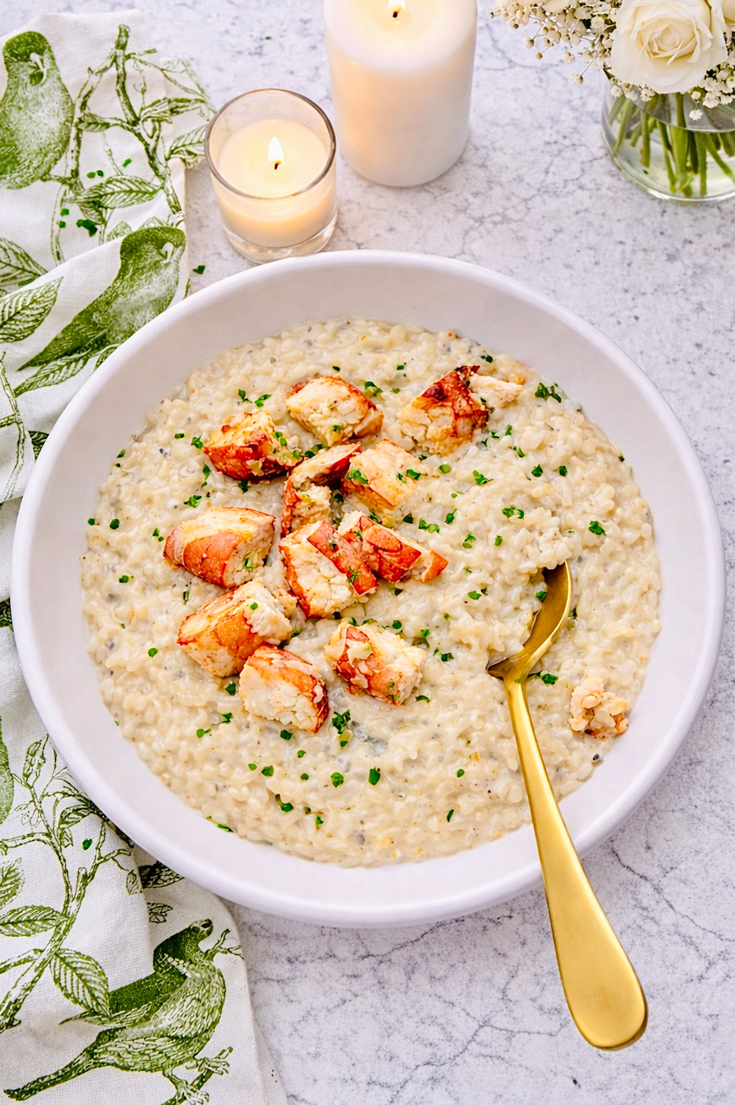 Overhead view of lobster risotto in a white bowl with lobster pieces, chives, and candles on a marble surface.