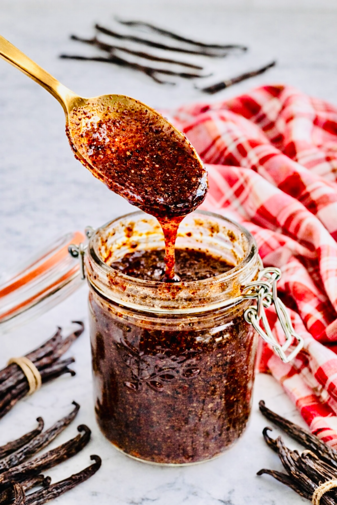Close-up of a gold spoon lifting thick homemade vanilla bean paste from a glass jar, with visible vanilla seeds and whole vanilla pods on a marble surface.