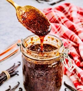 Close-up of a gold spoon lifting thick homemade vanilla bean paste from a glass jar, with visible vanilla seeds and whole vanilla pods on a marble surface.
