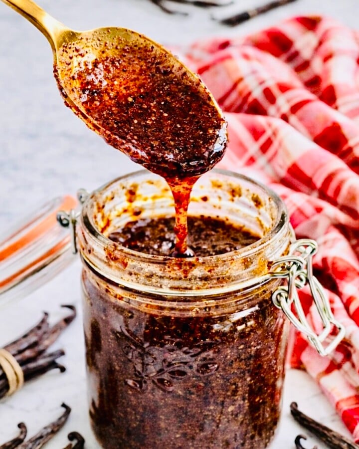Close-up of a gold spoon lifting thick homemade vanilla bean paste from a glass jar, with visible vanilla seeds and whole vanilla pods on a marble surface.