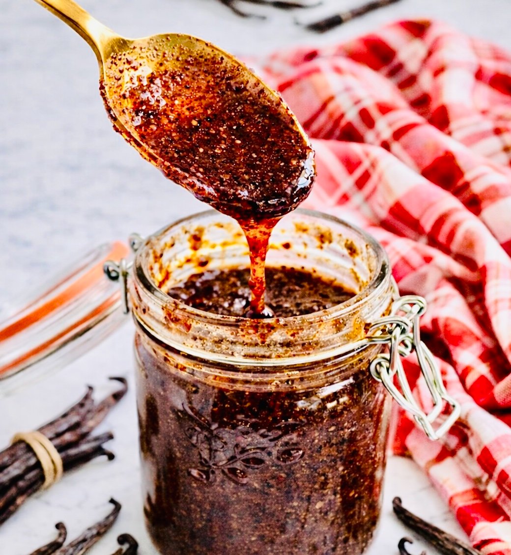 Close-up of a gold spoon lifting thick homemade vanilla bean paste from a glass jar, with visible vanilla seeds and whole vanilla pods on a marble surface.