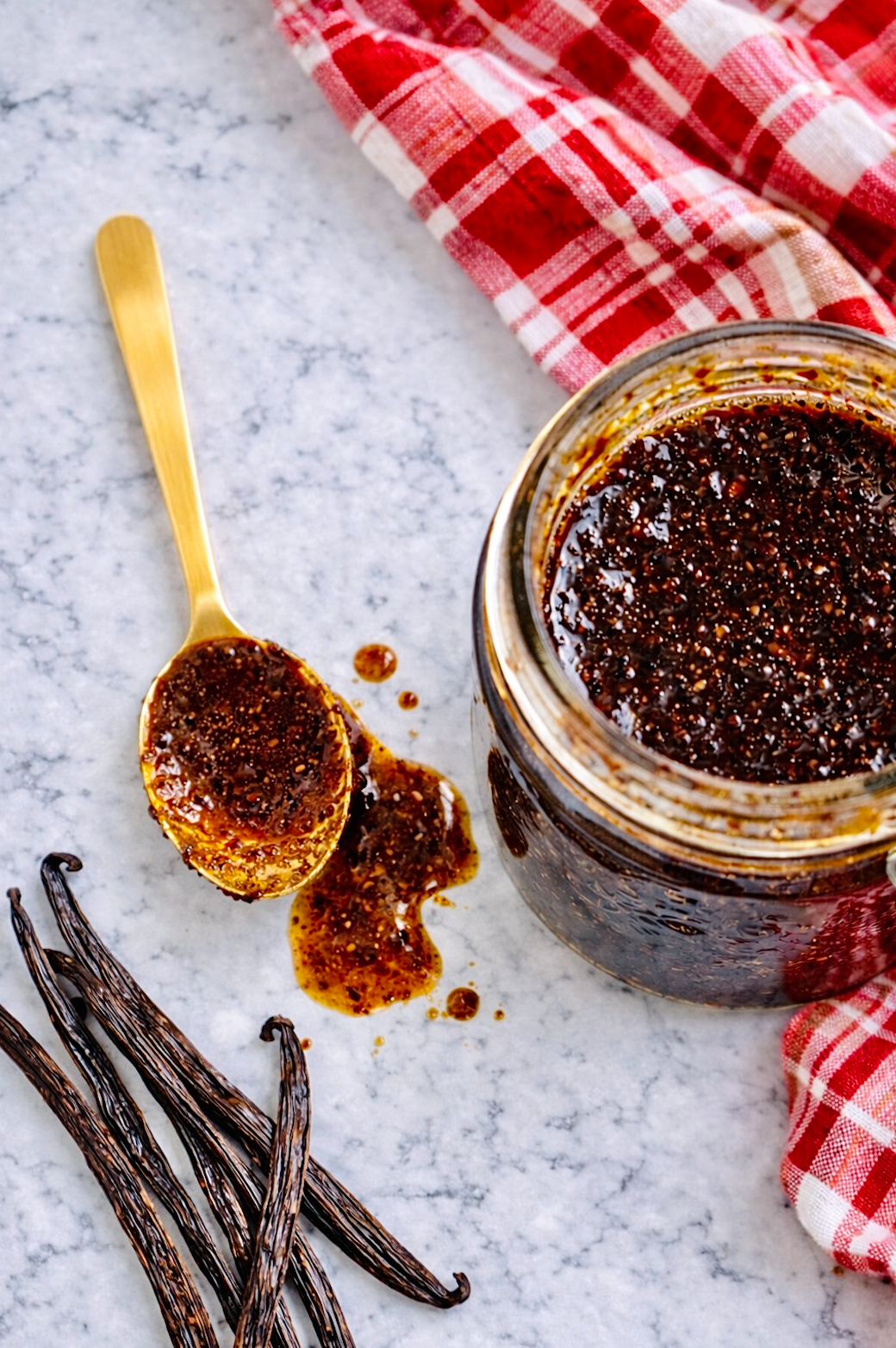 Overhead view of a jar of vanilla bean paste with a gold spoon resting beside it and whole vanilla bean pods scattered on a marble countertop.