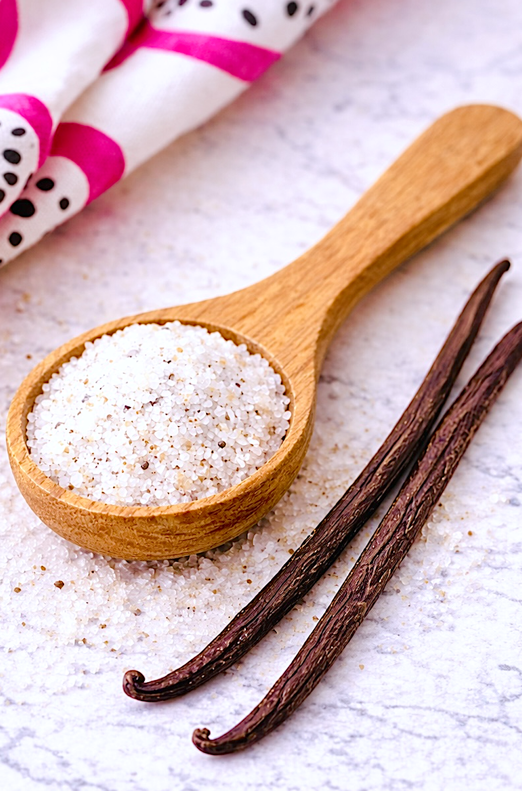 Close up of vanilla sugar in wooden spoon with whole vanilla beans on marble surface