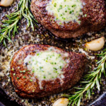 Overhead view of two ribeye steaks searing in a black cast iron skillet with melted garlic herb butter, rosemary sprigs, and coarse seasoning bubbling around the edges.