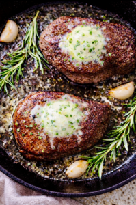 Overhead view of two ribeye steaks searing in a black cast iron skillet with melted garlic herb butter, rosemary sprigs, and coarse seasoning bubbling around the edges.