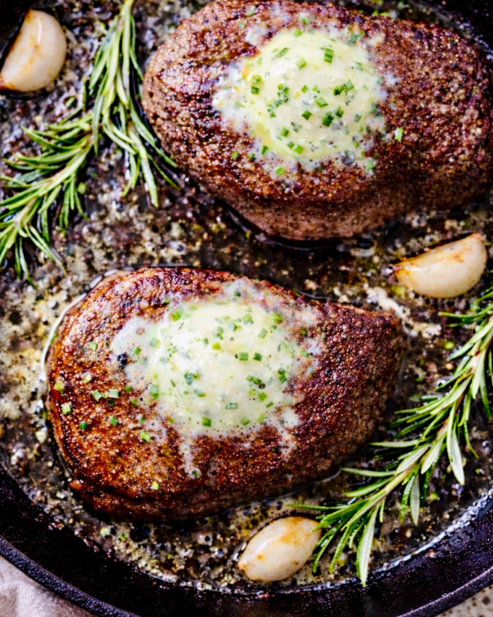 Overhead view of two ribeye steaks searing in a black cast iron skillet with melted garlic herb butter, rosemary sprigs, and coarse seasoning bubbling around the edges.