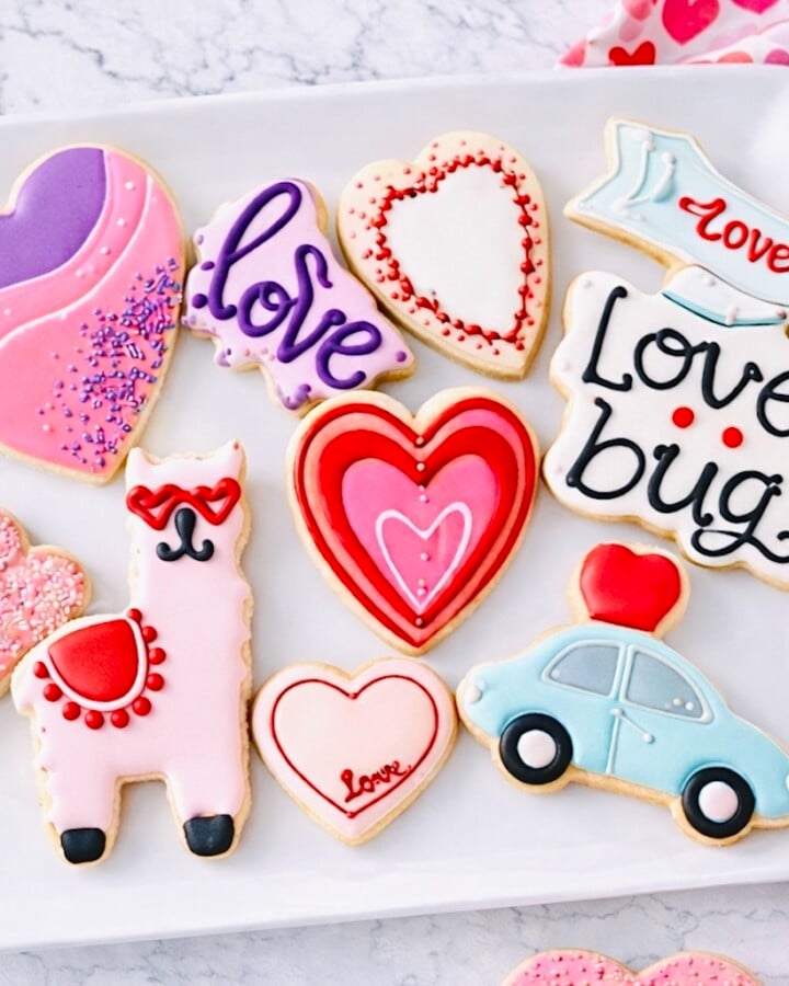 Decorated Valentine’s Day sugar cookies on a white plate with heart designs, royal icing details, and pink and red accents on a marble countertop.