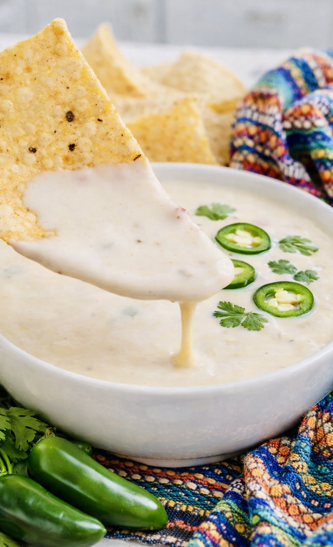 Creamy queso blanco dip in a white bowl garnished with sliced jalapeños and cilantro, served with tortilla chips on a colorful Mexican-style cloth.Creamy queso blanco dip in a white bowl garnished with sliced jalapeños and cilantro, served with tortilla chips on a colorful Mexican-style cloth.