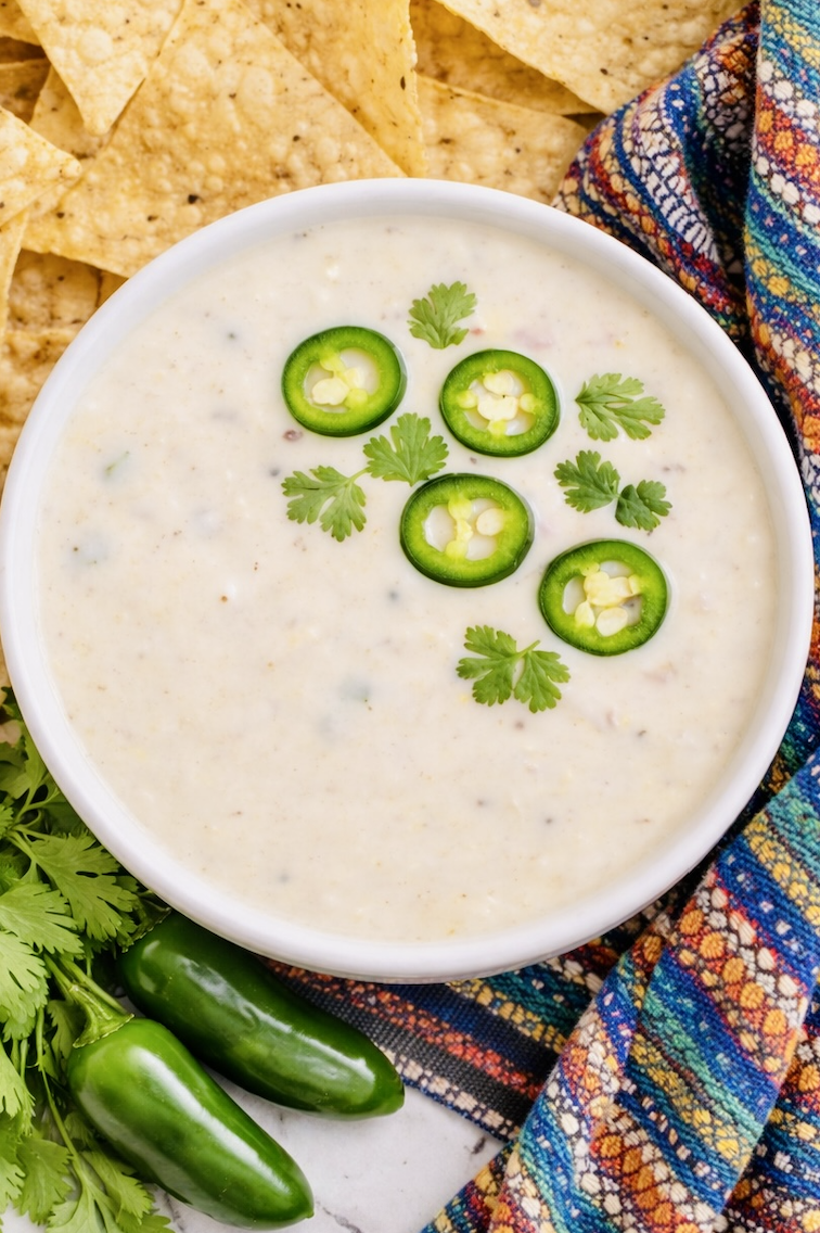 Overhead view of smooth white queso blanco topped with jalapeño slices and cilantro in a white bowl, surrounded by tortilla chips and fresh jalapeños.