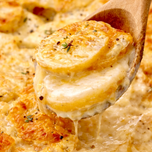 Wooden spoon lifting stacked slices of creamy scalloped potatoes from a golden baked casserole dish.