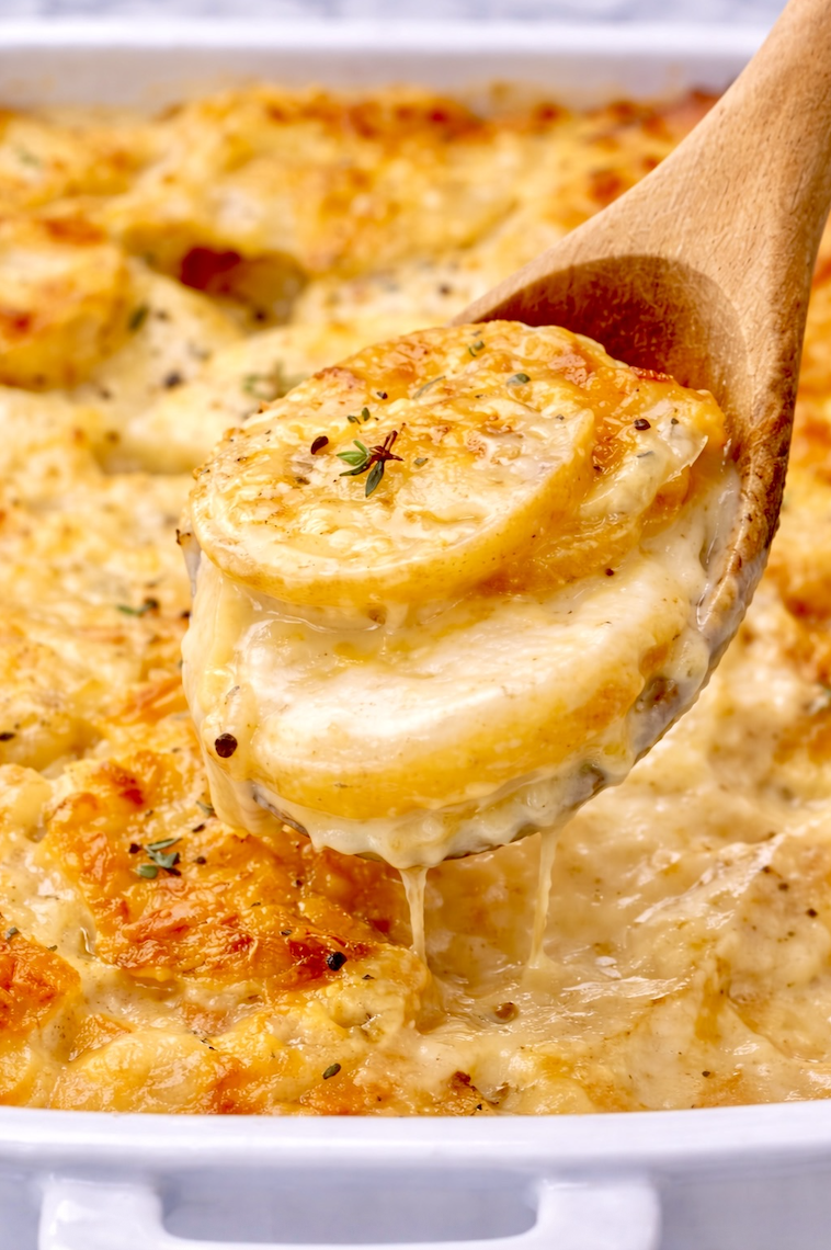 Wooden spoon lifting stacked slices of creamy scalloped potatoes from a golden baked casserole dish.