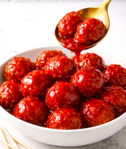 Spoon lifting grape jelly meatballs from white bowl with sauce dripping back into dish