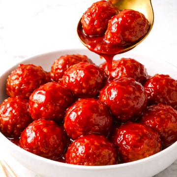 Spoon lifting grape jelly meatballs from white bowl with sauce dripping back into dish