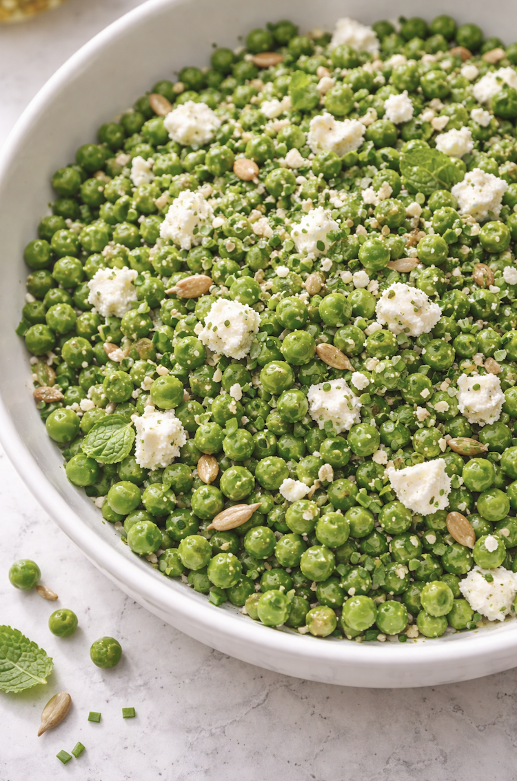 Overhead close-up of spring pea salad with green peas, feta crumbles, chopped chives, mint, and sunflower seeds in a white bowl