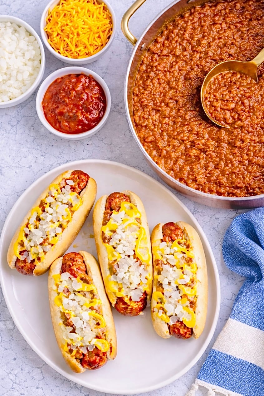 Overhead view of Coney Island hot dogs with chili sauce, mustard, chopped onions, and a pan of sauce