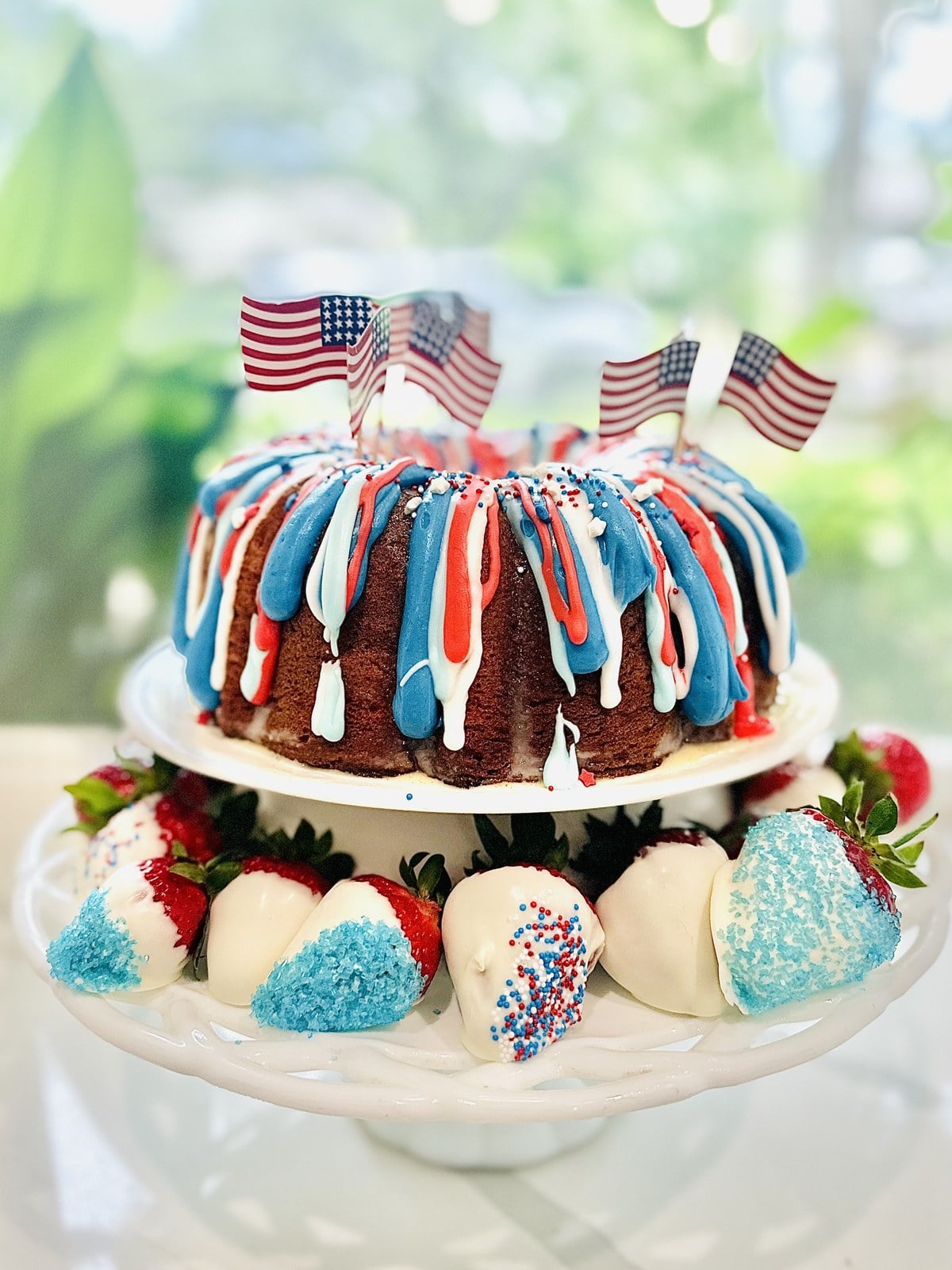 Side view of a patriotic Firecracker Bundt Cake with red, white, and blue glaze and decorated strawberries around the cake stand.