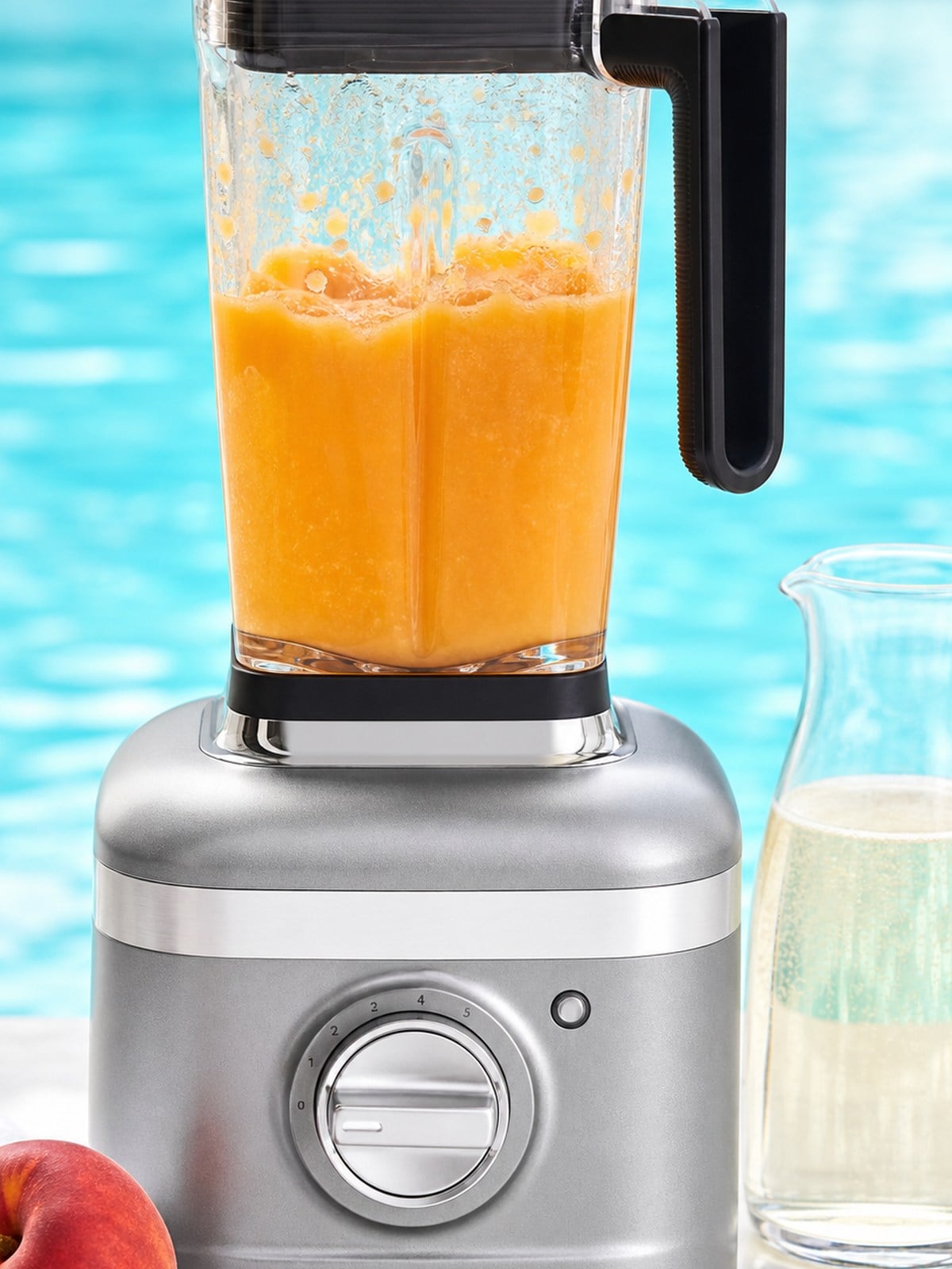 Smooth peach-orange frozen Bellini mixture in a silver KitchenAid K400-style blender with sparkling wine nearby.