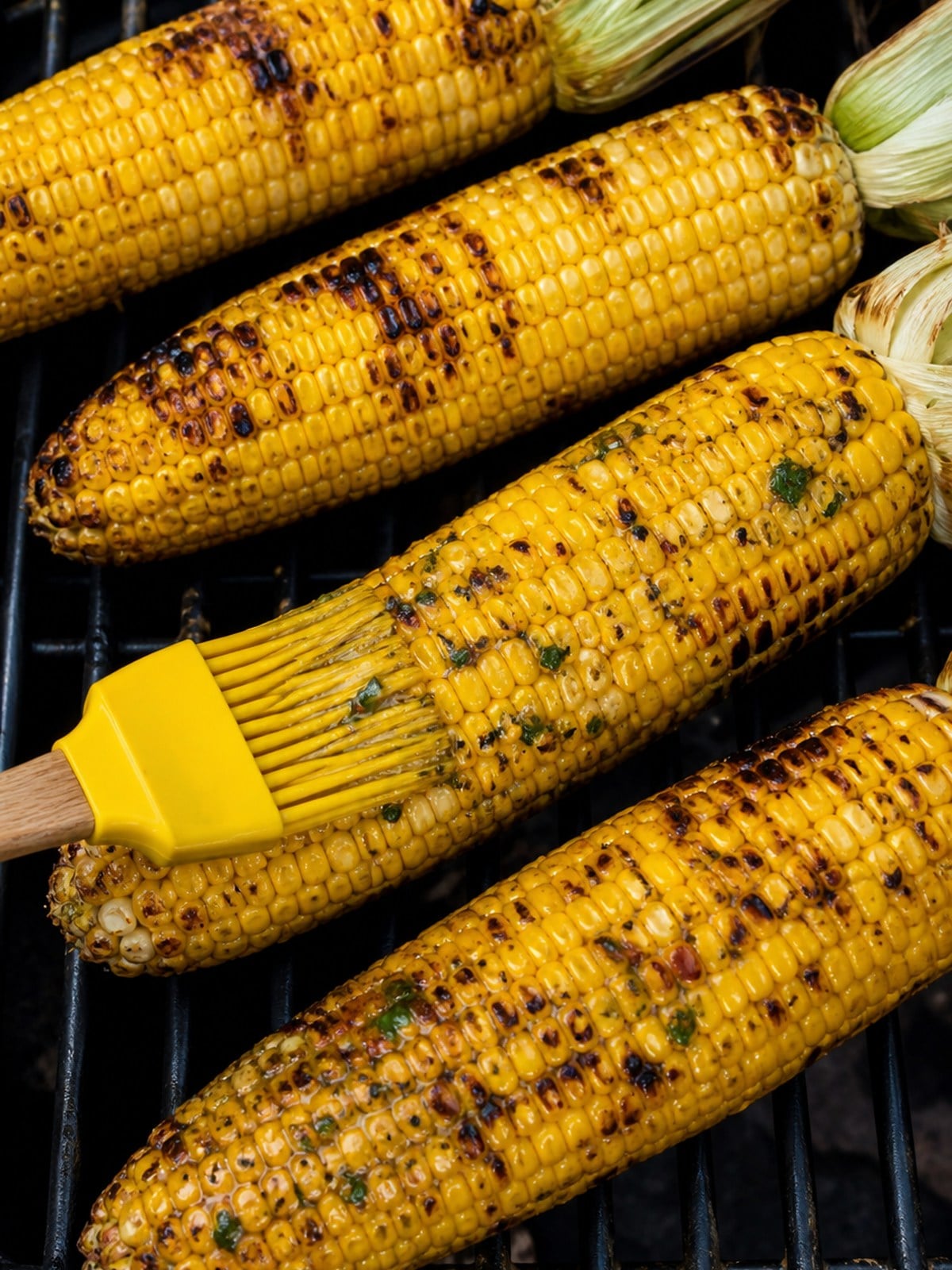 Fresh corn on a hot grill being brushed with melted garlic herb butter.