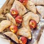 Southern fried peach pies coated in cinnamon sugar with fresh peaches on parchment.