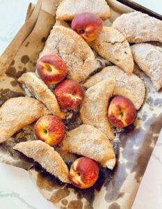 Southern fried peach pies coated in cinnamon sugar with fresh peaches on parchment.