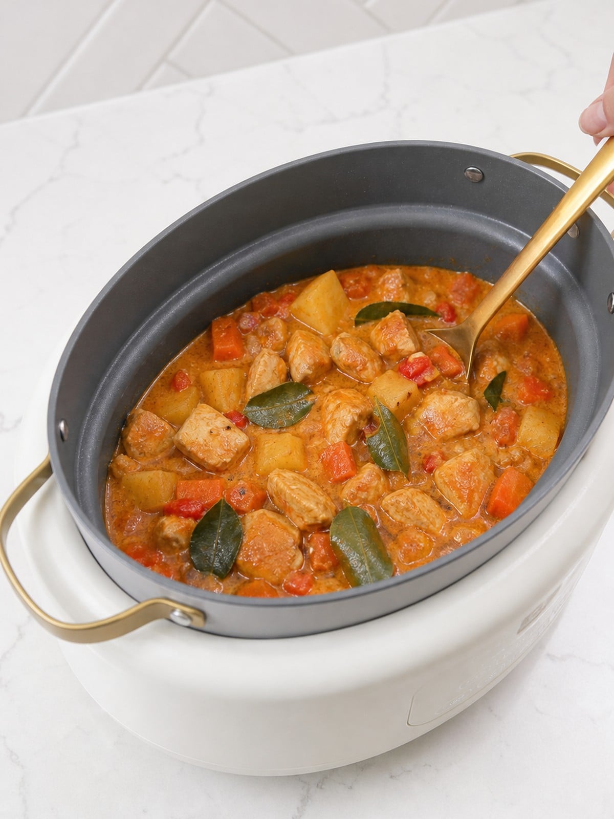 Massaman curry sauce stirred with chicken, vegetables, and kaffir lime leaves in a white slow cooker.
