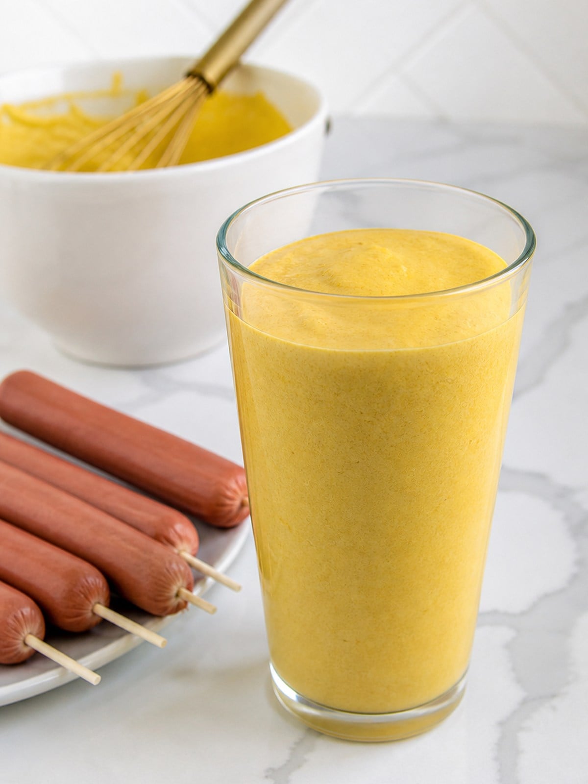 Tall glass filled with corn dog batter beside skewered hot dogs ready for dipping.