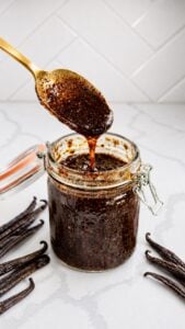 A gold spoon lifts thick, glossy homemade vanilla bean paste from a clear glass jar.