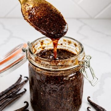 A gold spoon lifts thick, glossy homemade vanilla bean paste from a clear glass jar.