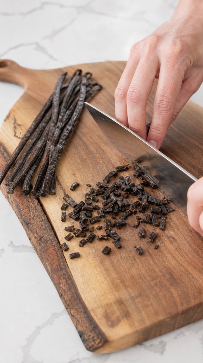 Whole vanilla beans being coarsely chopped on a wooden cutting board.