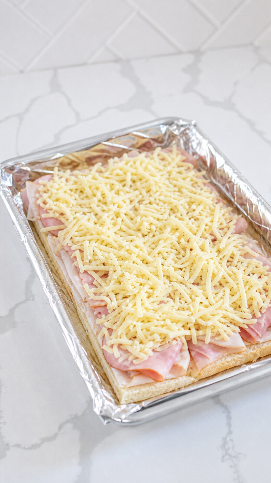 Monte Cristo sliders being assembled with ham turkey and grated cheese in a foil-lined baking dish.