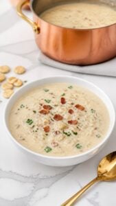 Finished easy New England clam chowder with small pieces of canned clams, peeled potatoes, bacon, and finely chopped parsley beside oyster crackers.