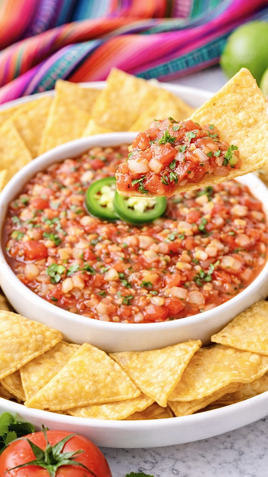 Tortilla chip lifting a scoop of chunky fresh tomato salsa from a white bowl.