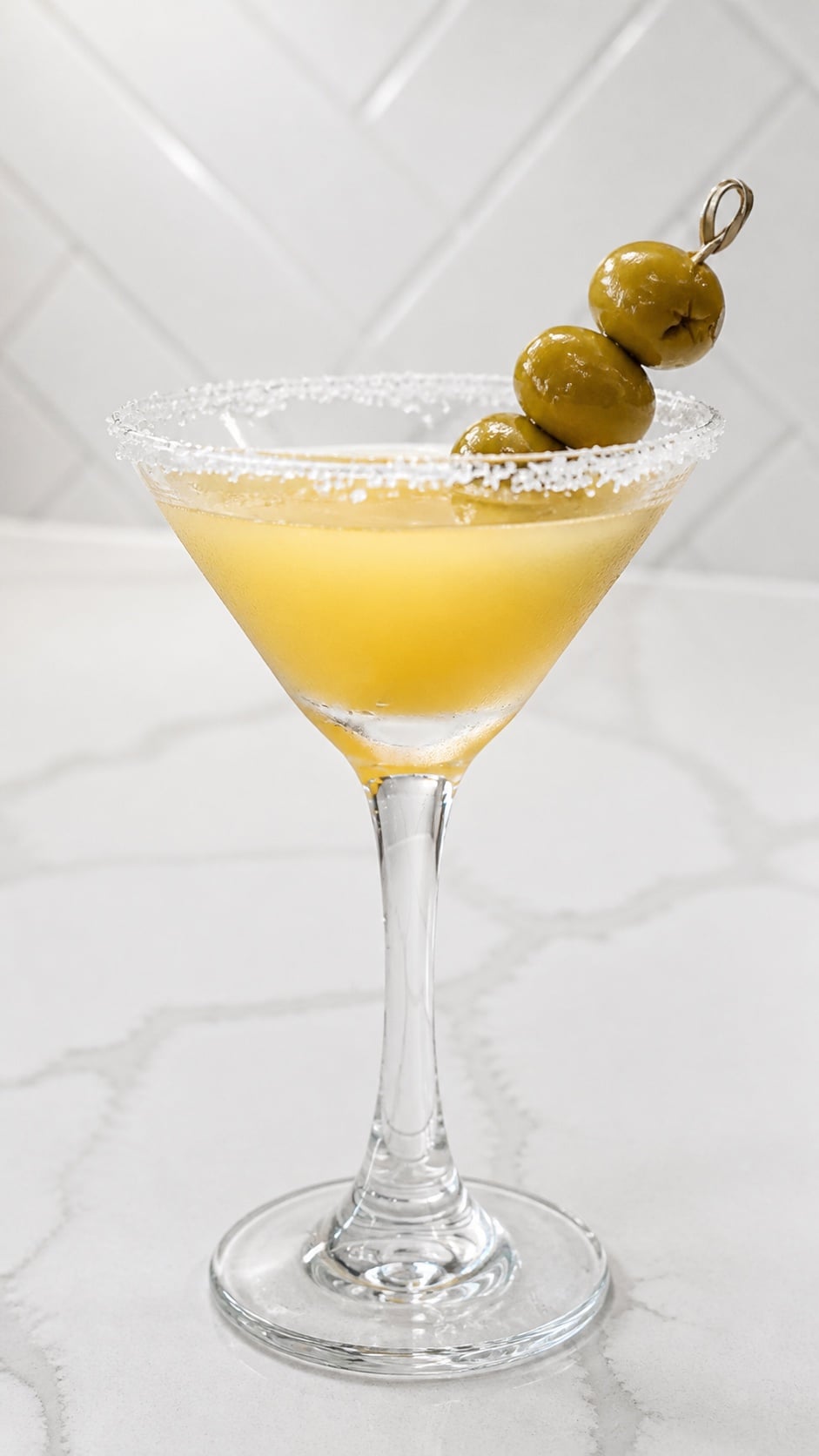 Close-up Mexican martini showing the coarse salt rim, condensation, and green olive garnish.