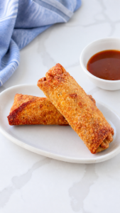 Two crisp homemade pork egg rolls on an ivory platter with sweet and sour sauce and a softly bunched blue striped linen.