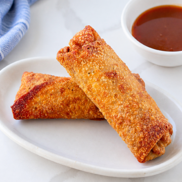 Two crisp homemade pork egg rolls on an ivory platter with sweet and sour sauce and a softly bunched blue striped linen.